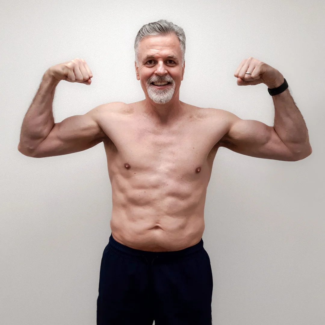 Smiling middle-aged man flexing his muscles shirtless against a plain white background.