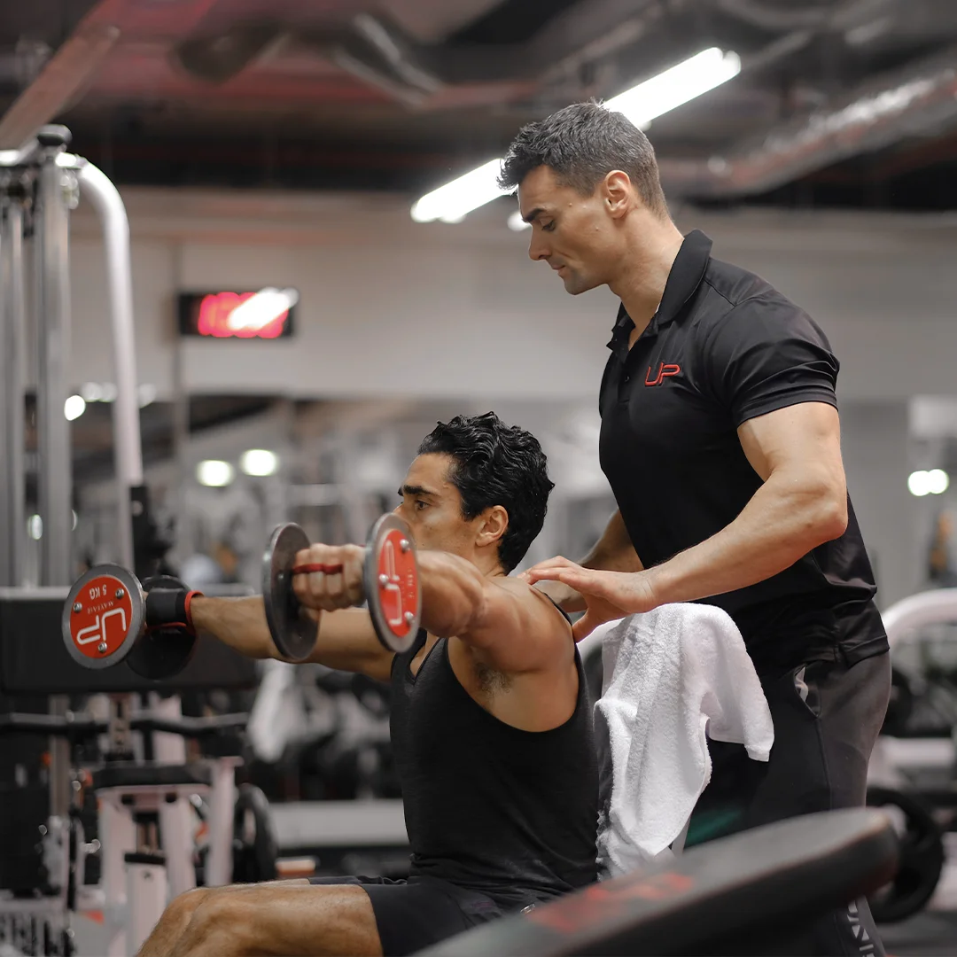 A fitness trainer assists a man lifting dumbbells during a shoulder workout in a gym.