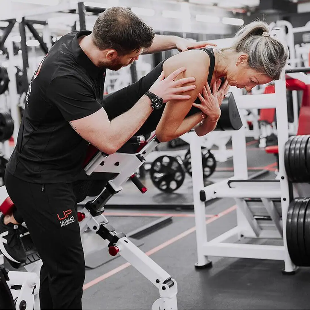 Fitness trainer assisting an older woman performing an inclined back extension exercise on a gym machine.