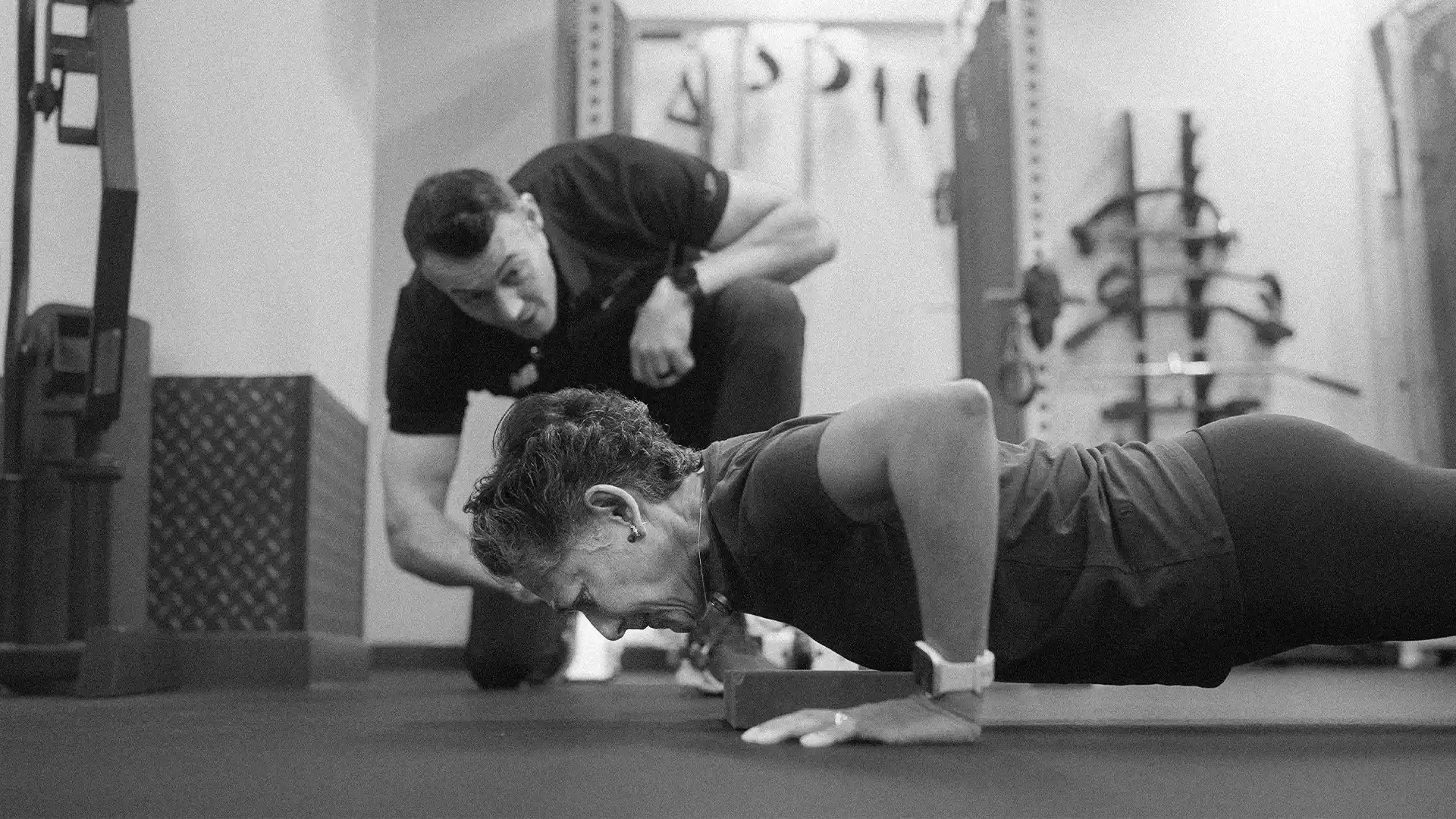 Older woman doing a push-up on the floor while a male trainer watches and encourages her in a gym.