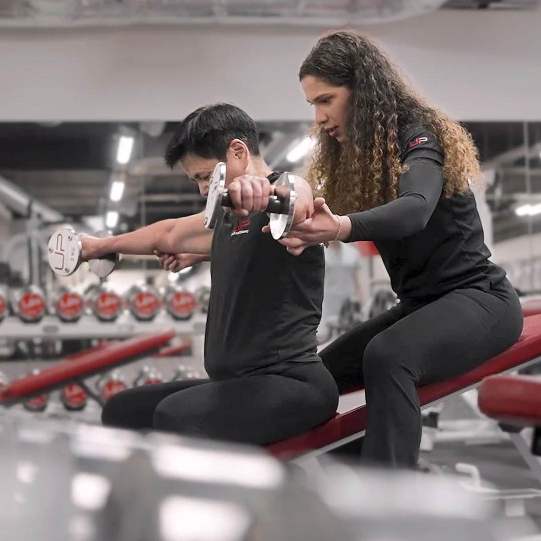 Trainer assisting a man lifting dumbbells while seated on a bench in a gym.