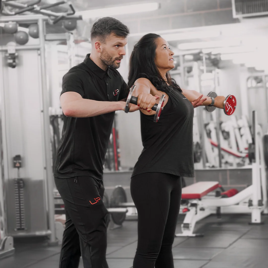Female trainer assisting woman with dumbbell lateral raises in a gym setting.