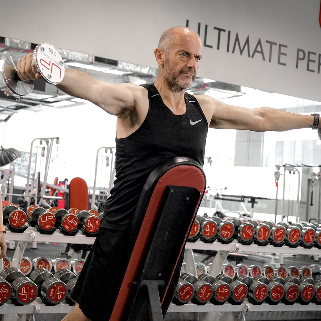 Man in black tank top lifting dumbbells with arms extended in a gym with rows of red and silver weights behind him.