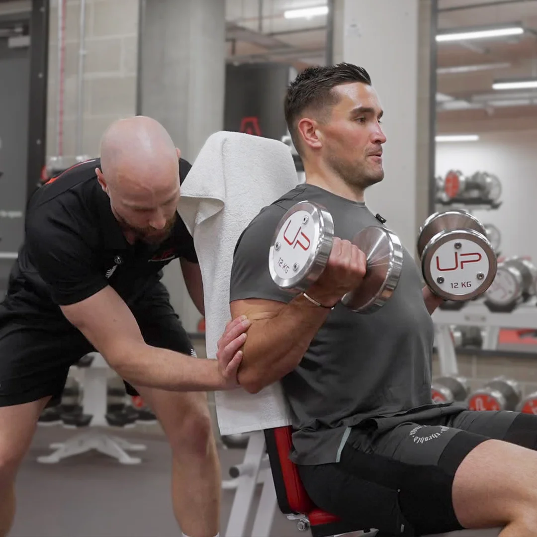 Trainer assists a man performing seated dumbbell curls with 12 kg weights in a gym.
