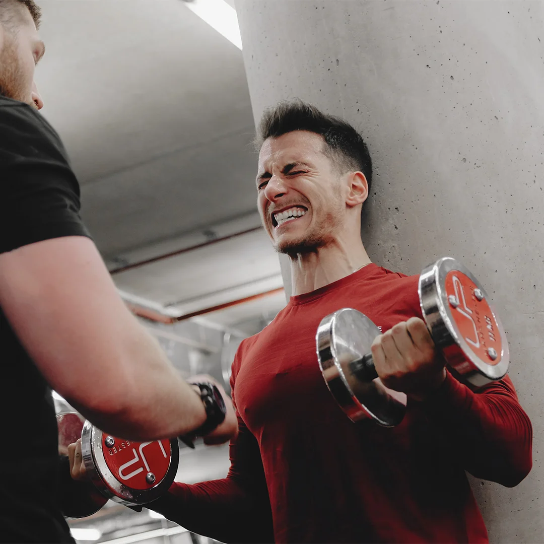 Man in a red shirt lifting dumbbells with a strained expression while a trainer supports him in a gym.