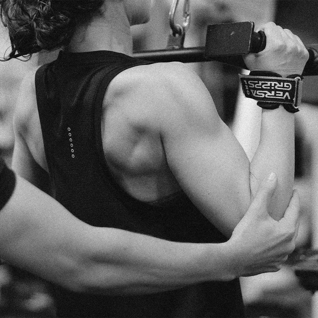 Close-up of a person lifting a barbell with wrist straps, assisted by another person's hand supporting their arm.