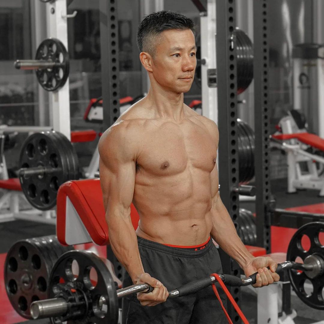 Muscular man performing barbell curls in a gym with weight plates in the background.