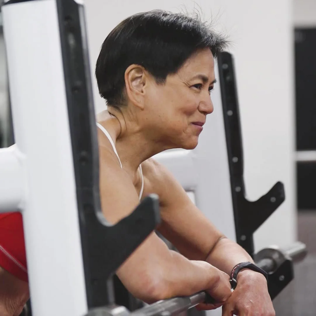 Smiling middle-aged woman resting on a barbell rack in a gym.