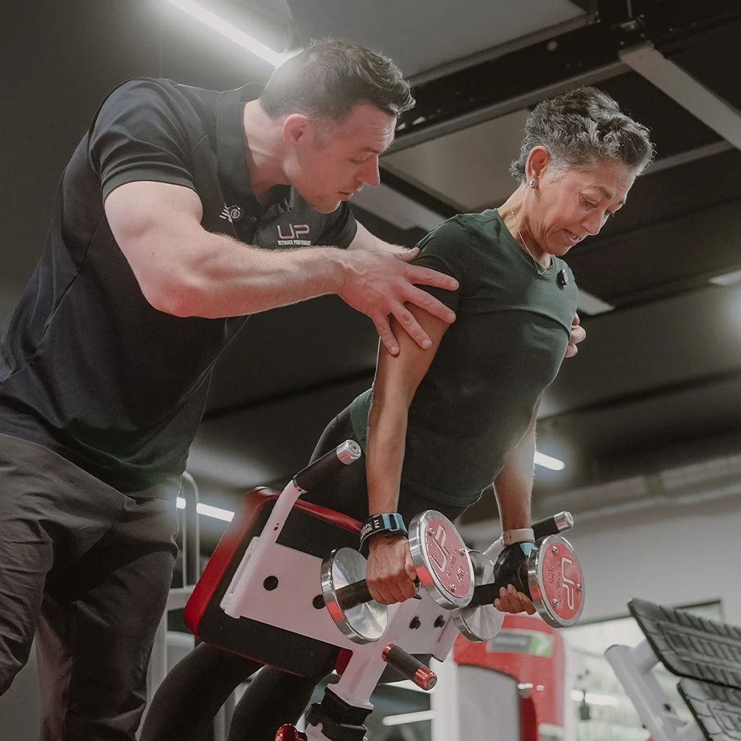 Fitness trainer assisting a woman holding weights while performing an inclined exercise on a gym bench.