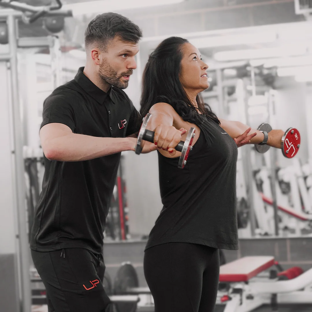 Fitness trainer assisting woman lifting dumbbells in a gym during exercise.