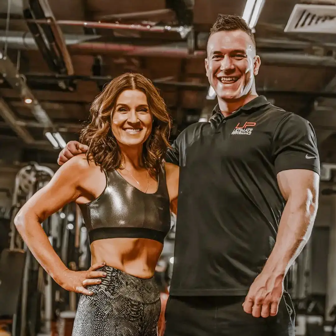 Smiling fit woman in sports bra and patterned leggings standing next to muscular man in black Ultimate Performance polo in a gym.