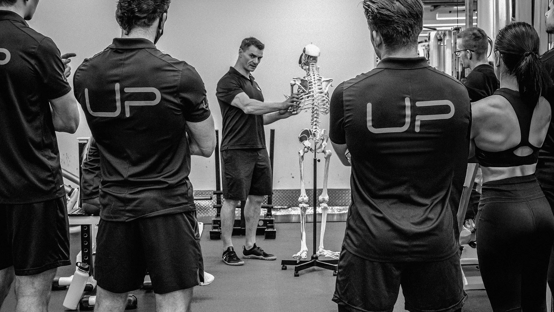 Fitness instructor demonstrating human skeletal anatomy to a group of trainees in a gym setting.