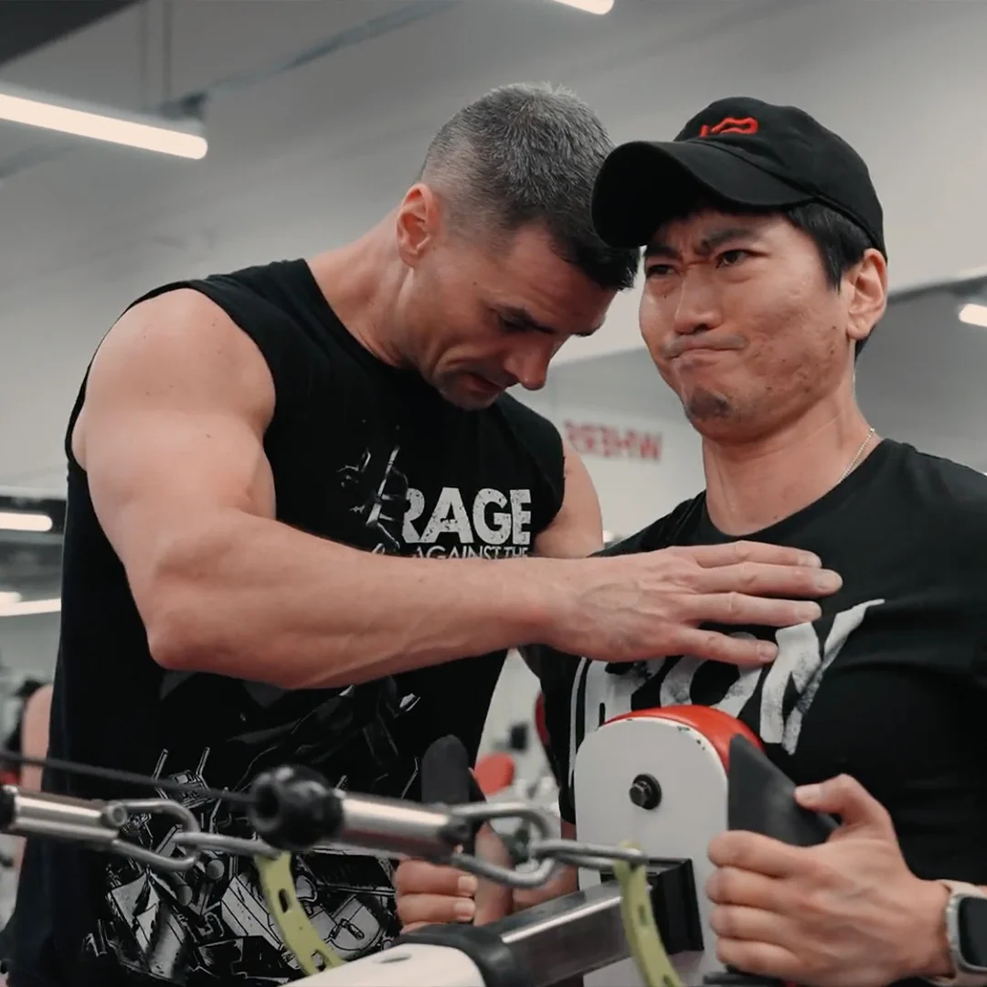 Trainer adjusting a fitness machine while instructing a focused man wearing a black cap in a gym.