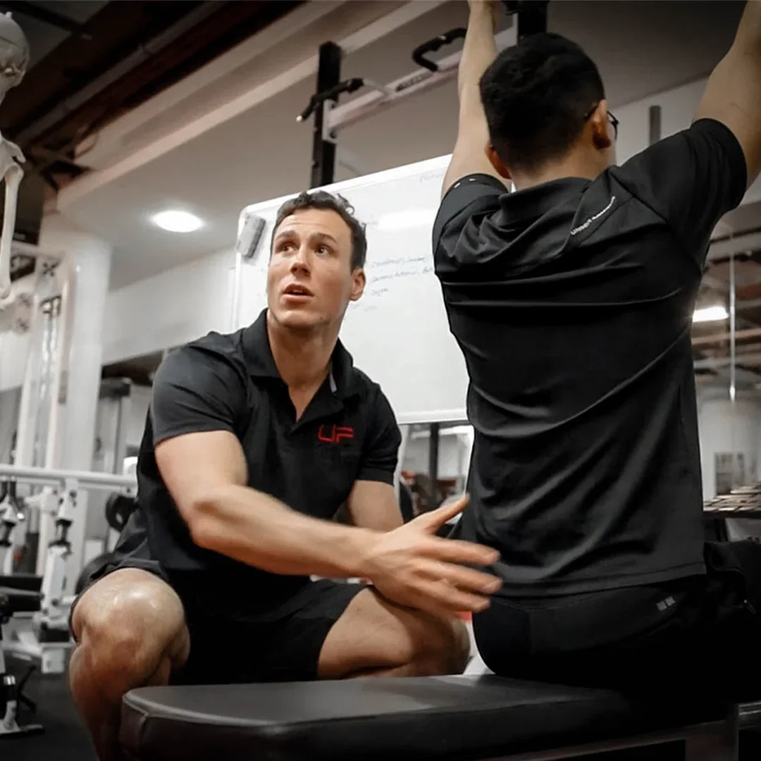 Personal trainer coaching client performing a seated exercise with hands raised at a gym.