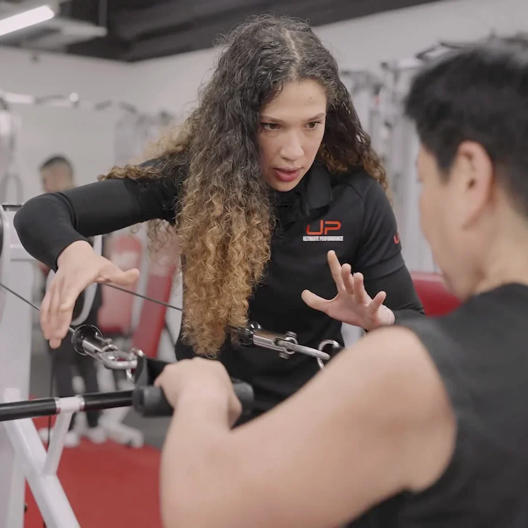 Female fitness trainer with curly hair instructing a man using gym cable machine.