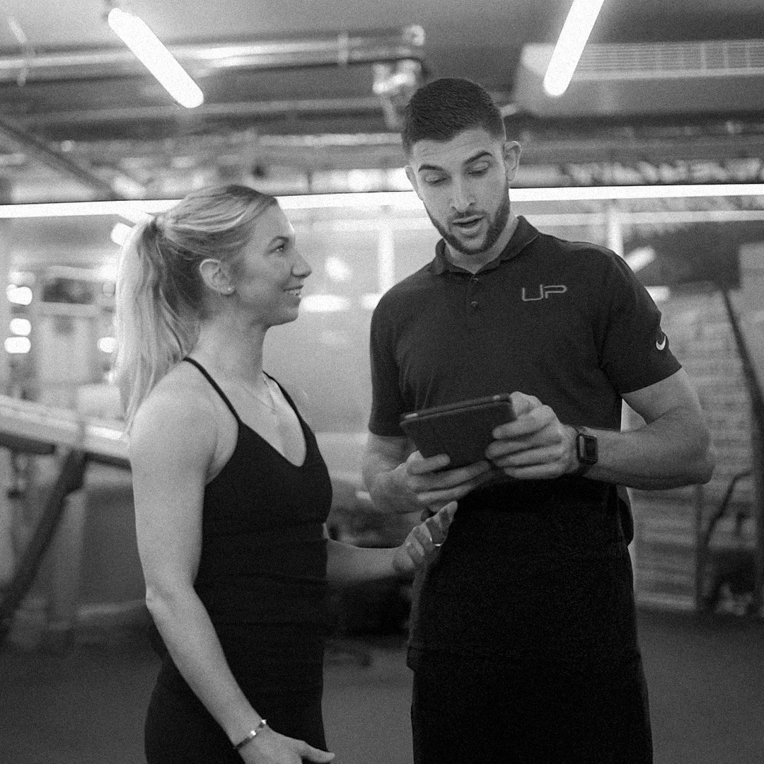 Female fitness client talking with a male trainer holding a tablet in a gym.