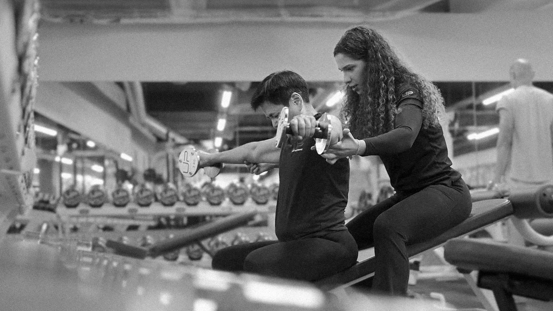 A fitness trainer assists a person lifting dumbbells while seated on a bench in a gym.