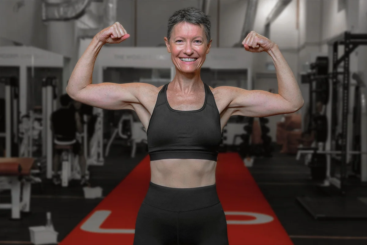Smiling fit woman in black workout attire flexing her arm muscles in a gym.