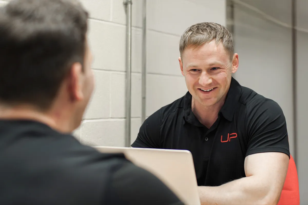Two men in black shirts smiling and talking across a table with a laptop in a modern office setting.