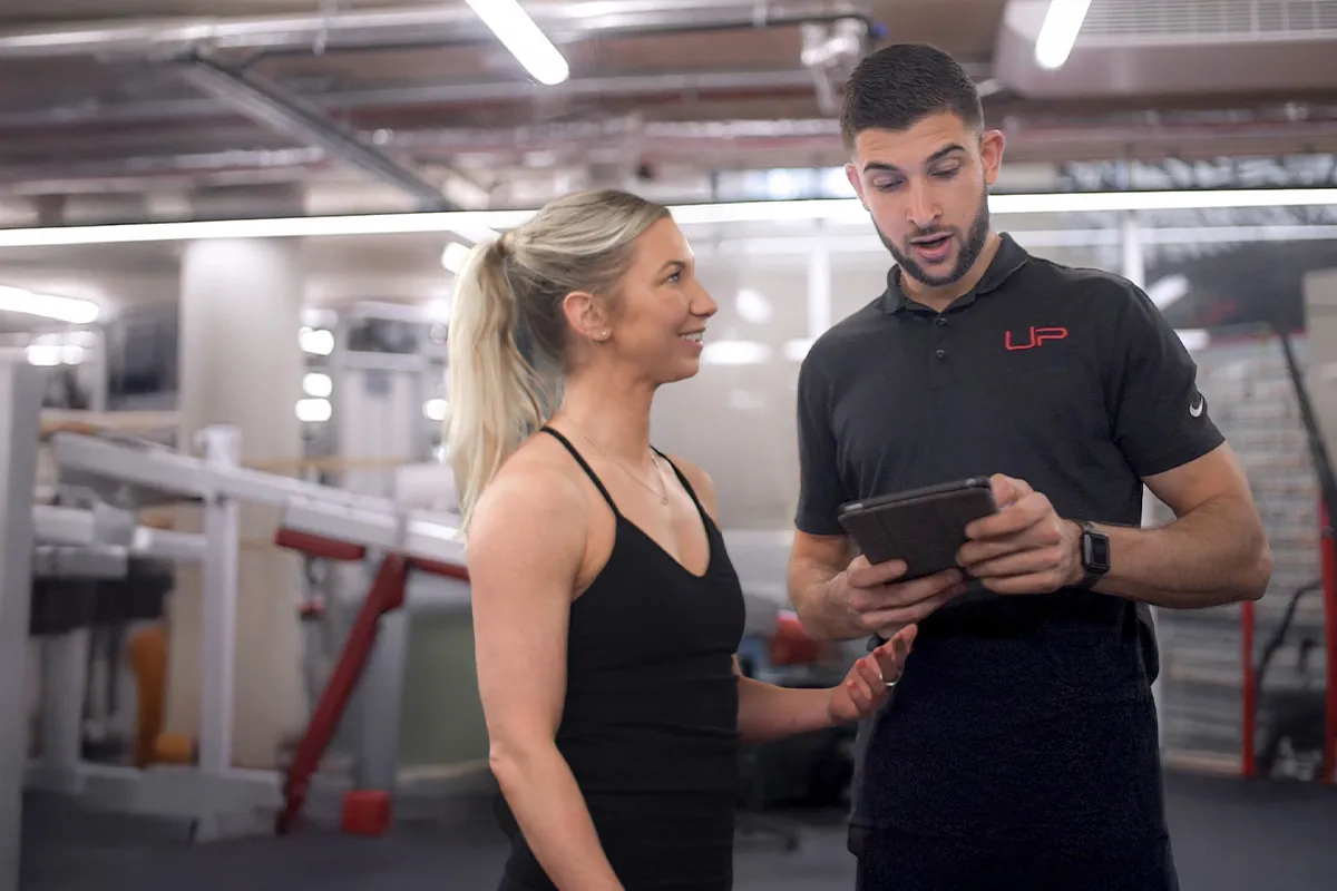 A female gym member in black workout clothes talks with a male trainer holding a tablet in a gym setting.