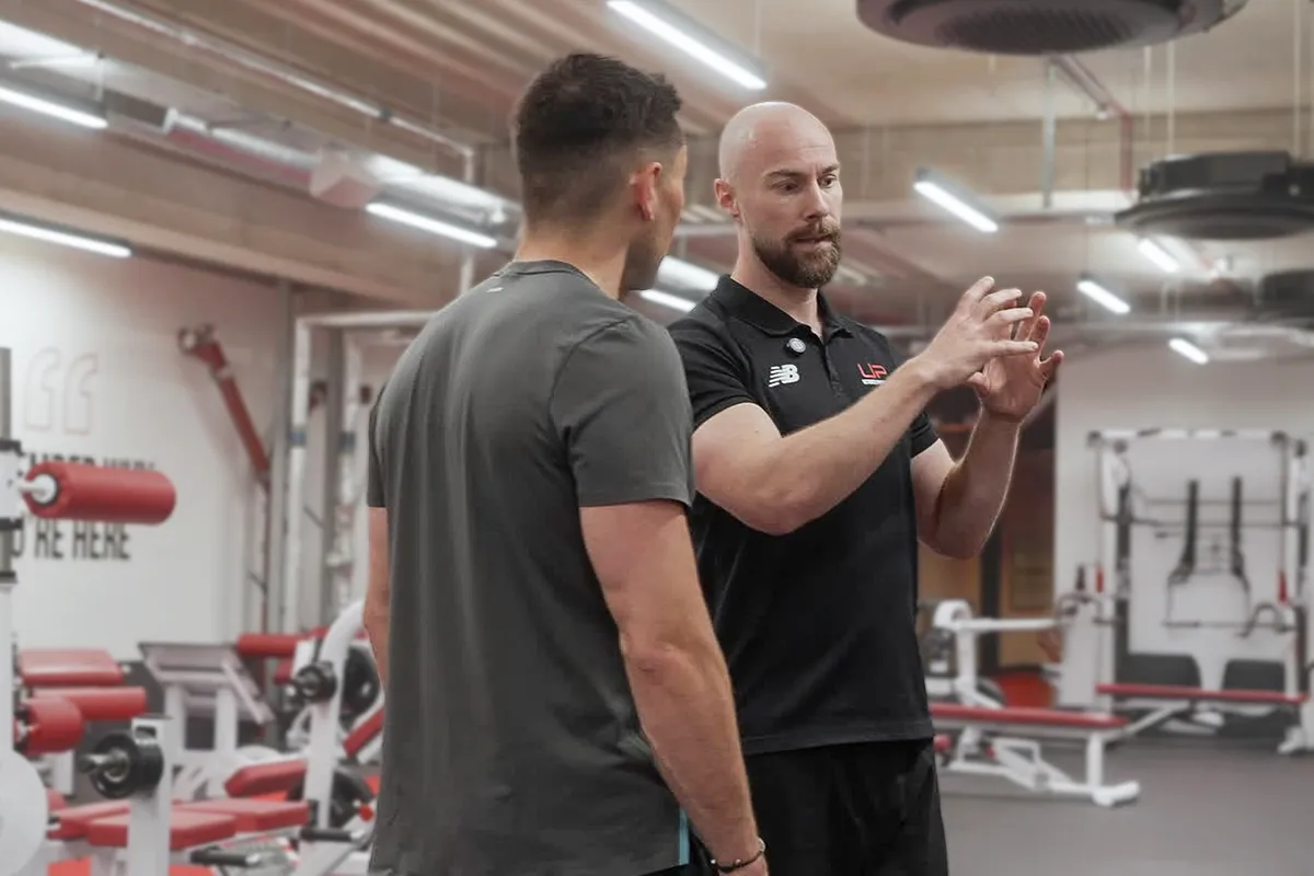 A fitness trainer in black explaining a workout technique to a man in grey in a gym setting.