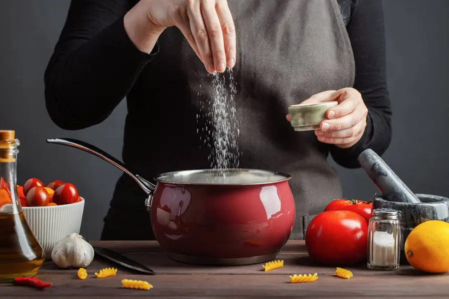Person sprinkling salt into a red saucepan on a wooden table with tomatoes, garlic, pasta, and cooking ingredients around.