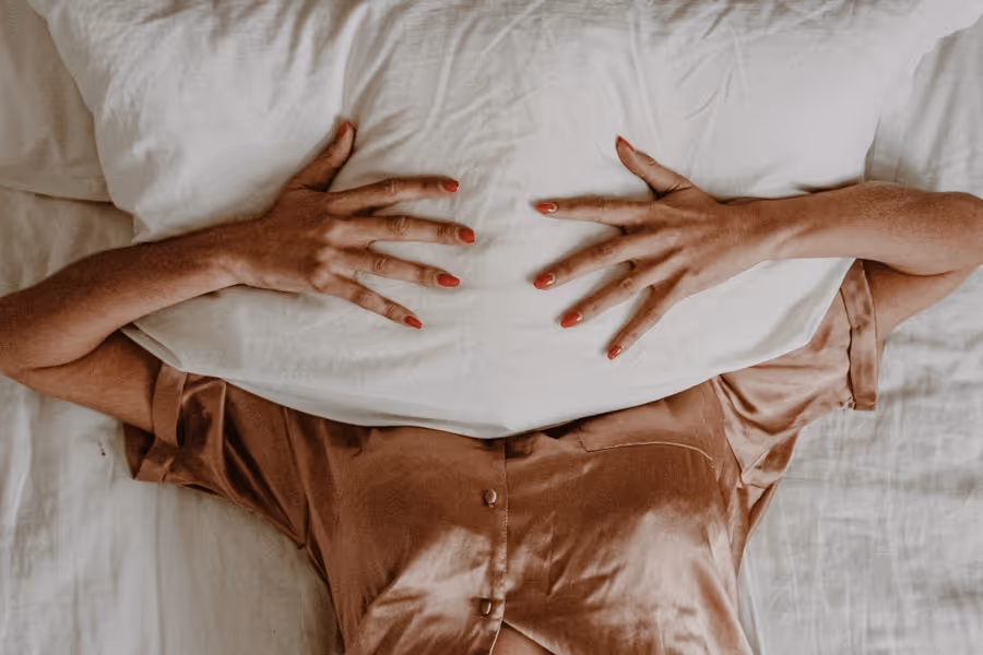 Person lying on a bed wearing a satin brown shirt, covering their face with a white pillow and hands with red nail polish visible.
