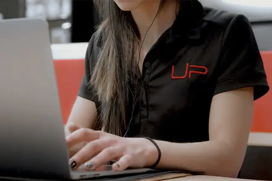 Person with long hair wearing a black shirt with red 'UP' logo typing on a laptop.