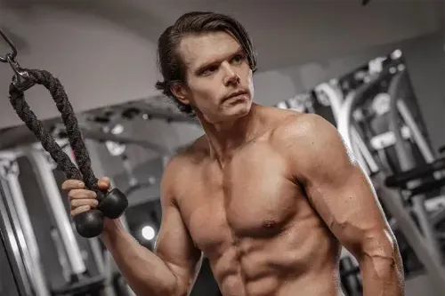 Muscular shirtless man exercising with gym cable machine attachment in fitness center.