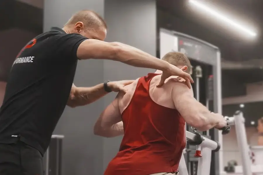 Trainer assisting a man in a red tank top with back exercise on a machine in a gym.