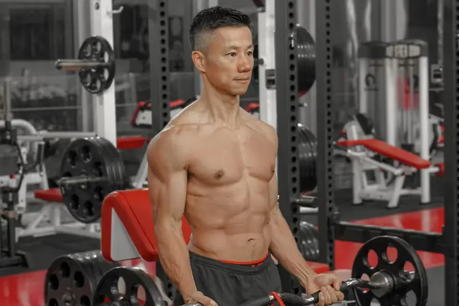 Muscular man with a bare torso lifting weights in a gym setting with red and black equipment.