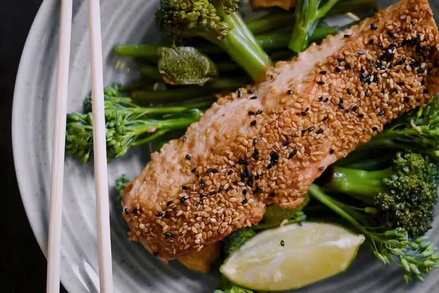 Sesame-crusted salmon fillet served on a bed of steamed broccoli with a lime wedge and chopsticks on the side.