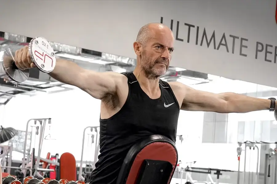 Man in black sleeveless shirt lifting dumbbells with arms extended sideways in a gym.