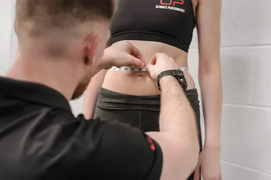 Trainer measuring a woman's waist with a tape measure in a fitness setting.