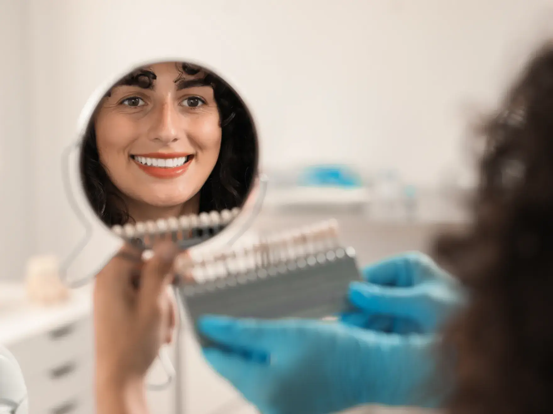 Woman smiling at her reflection in a handheld mirror while a gloved dental professional holds a dental shade guide.