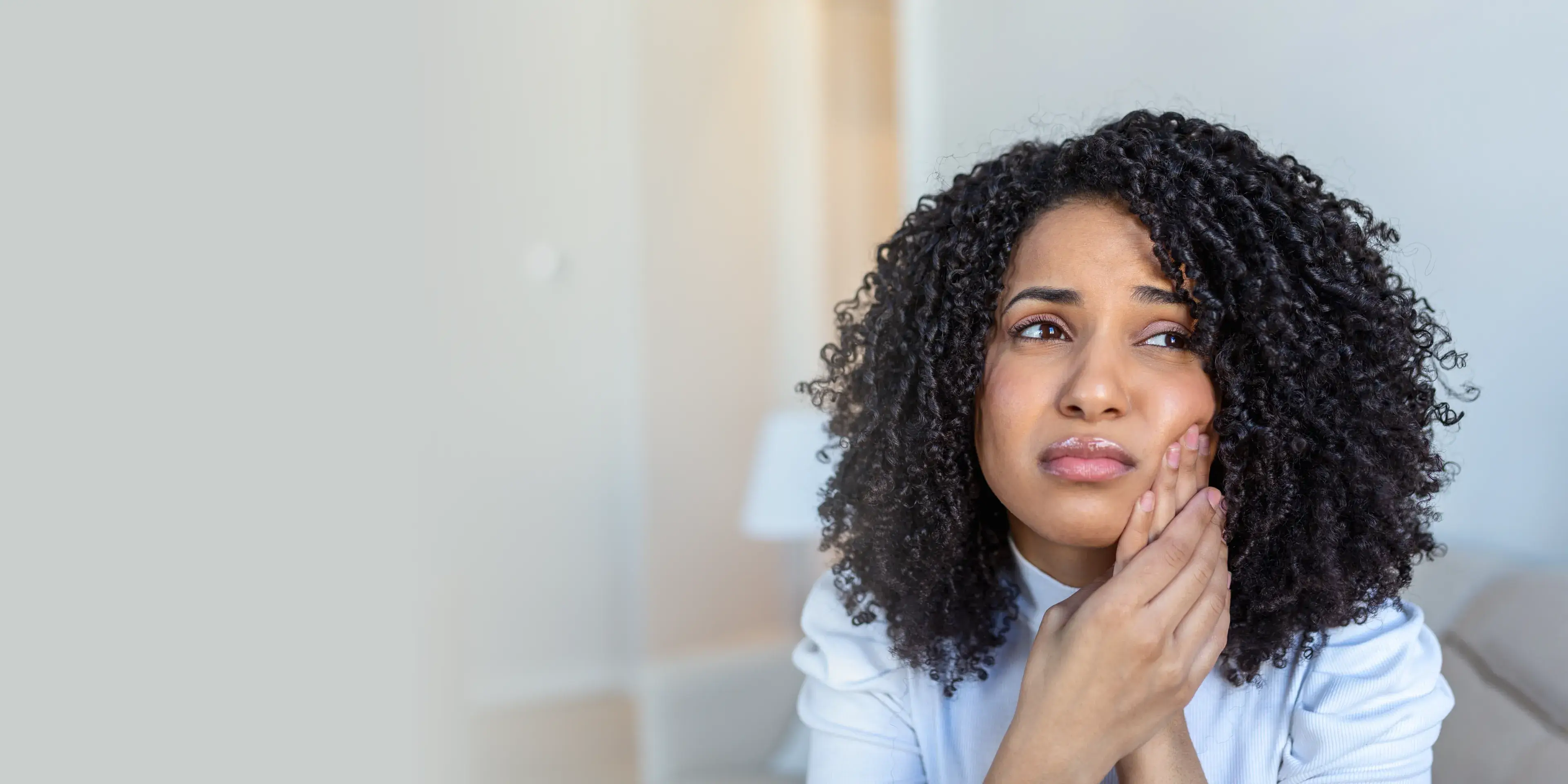 Woman with curly hair holding her cheek, showing signs of toothache or dental pain.