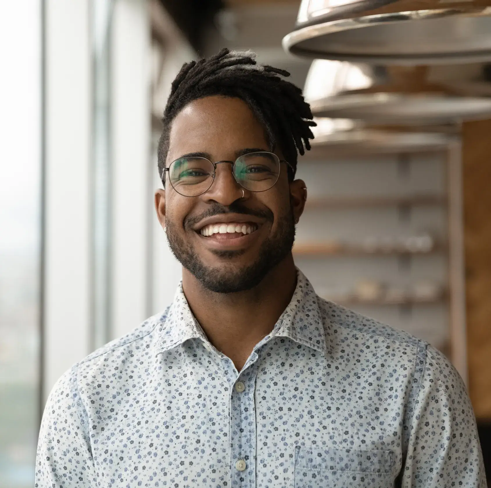 Smiling man with glasses and dreadlocks wearing a white patterned shirt indoors near a window.