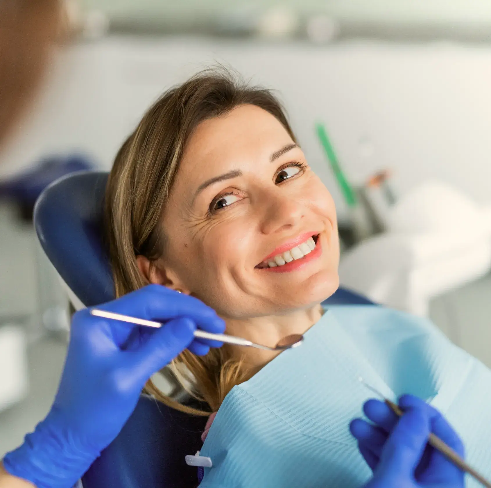 Smiling woman reclining in a dental chair with dentist hands in blue gloves holding dental tools near her mouth.
