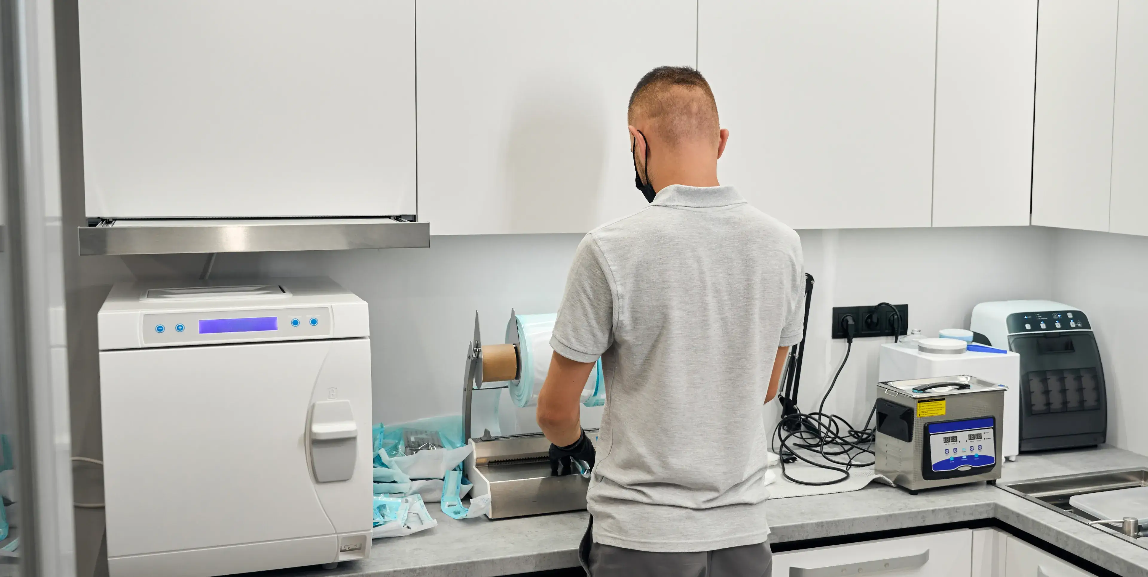 Man wearing black gloves and a mask preparing sterilization pouches in a clean dental sterilization room.