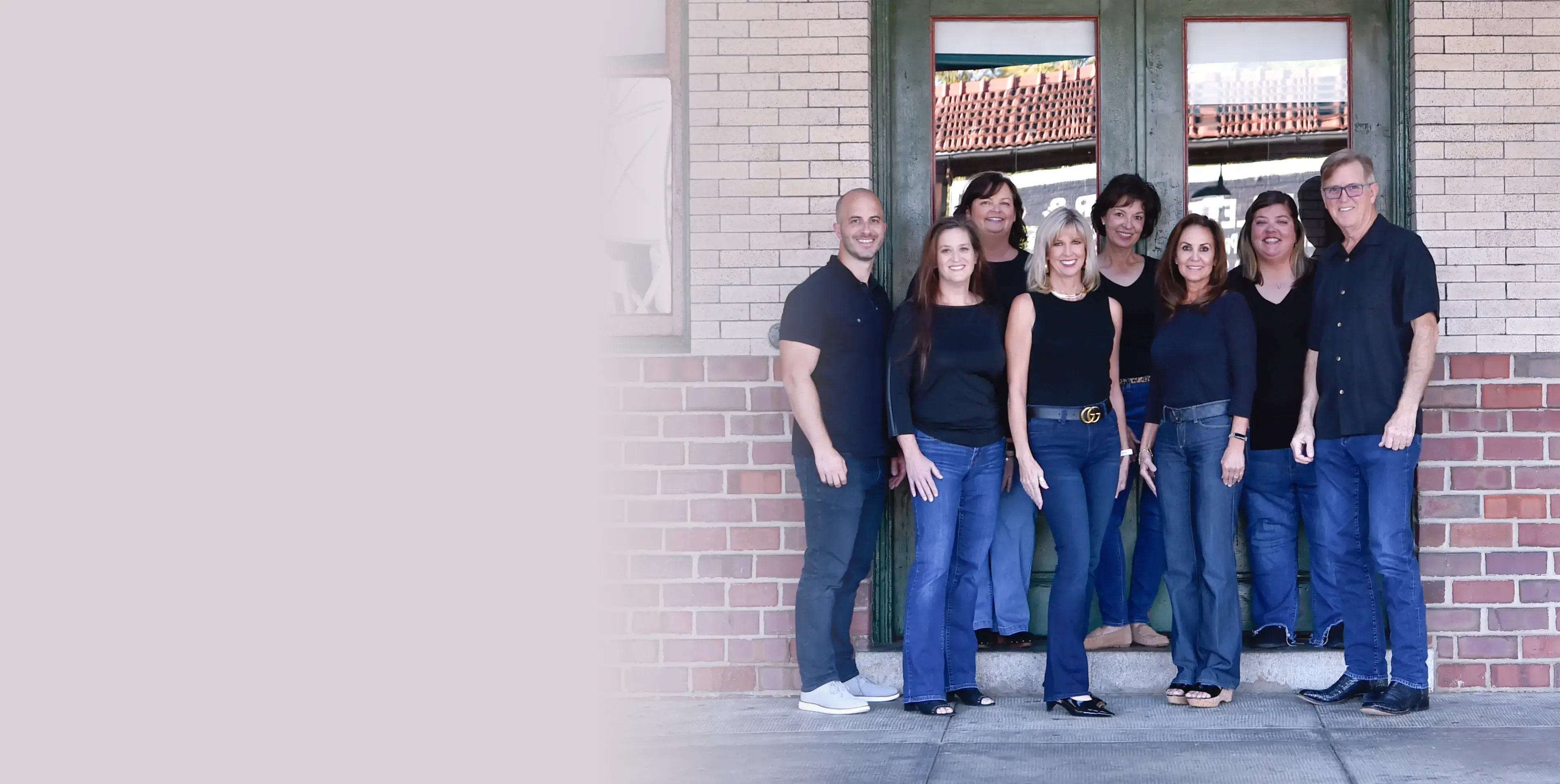Group of eight adults standing together in front of a green door with brick walls, all smiling and dressed in casual dark tops and jeans.