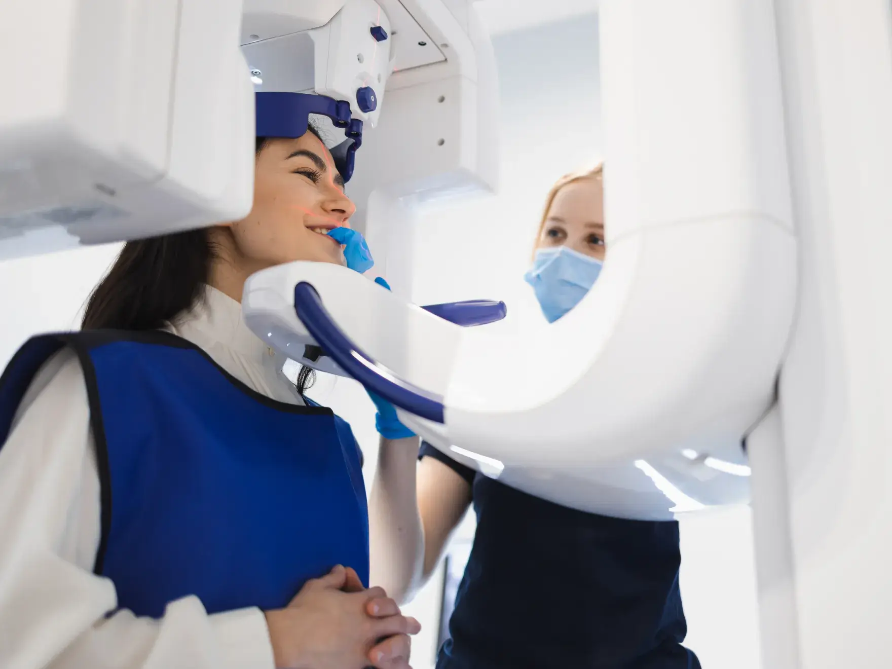 Dental technician wearing a mask positioning a woman in a protective blue apron for a dental X-ray scan.