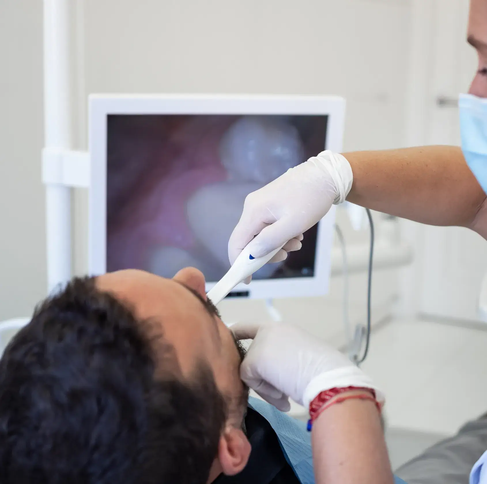 Dentist wearing gloves using an intraoral camera inside a male patient's mouth with the image displayed on a monitor.