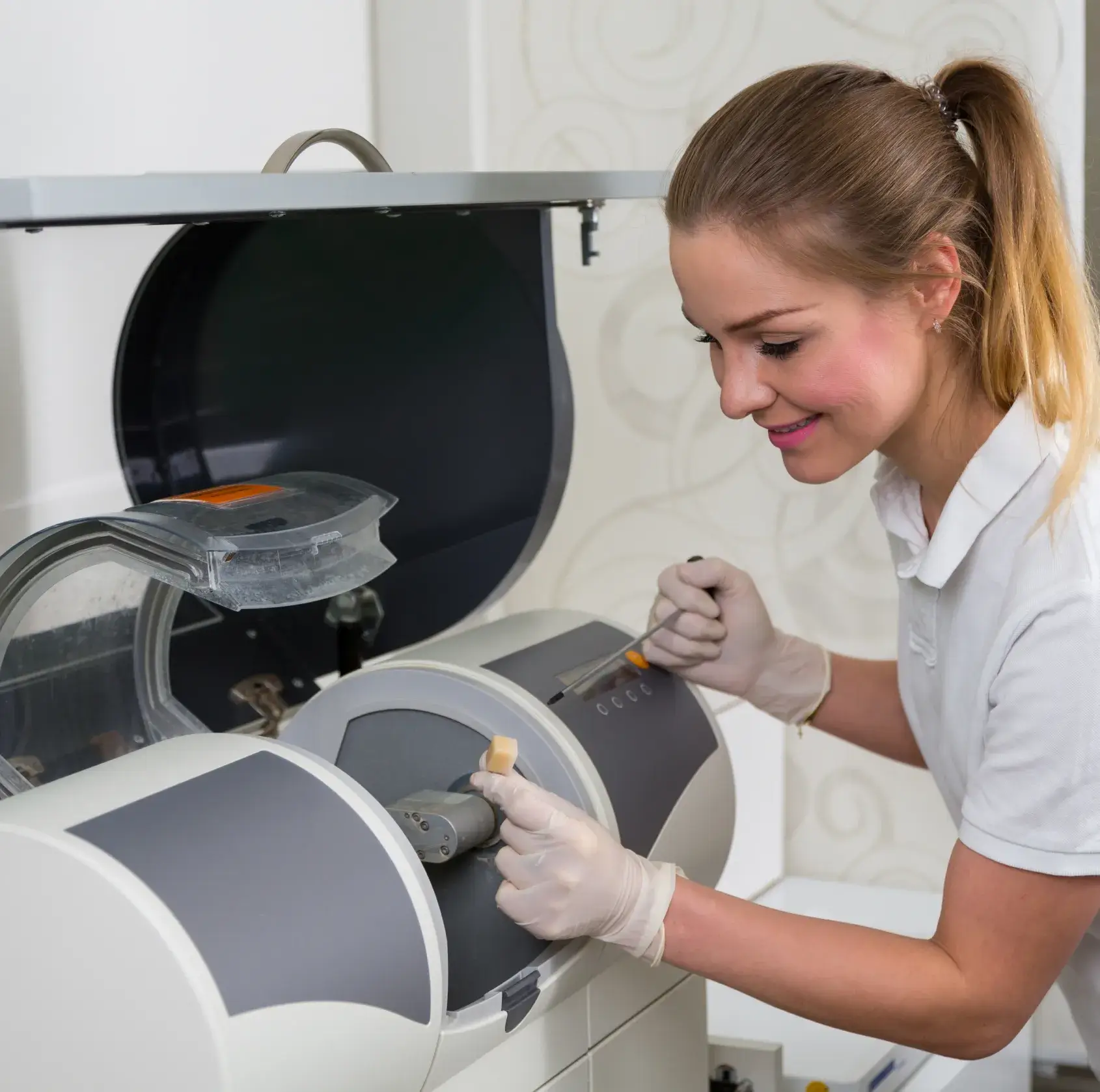 Dentist wearing gloves operating a dental milling machine, holding a dental ceramic block and a tool.