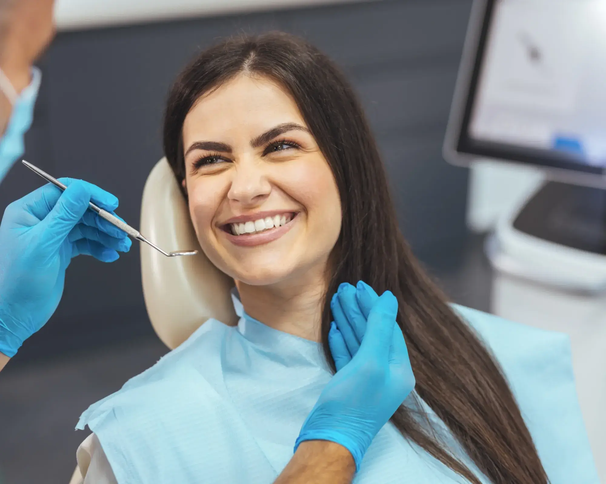 Smiling woman in a dental chair receiving a checkup from a dentist wearing blue gloves holding a dental mirror.