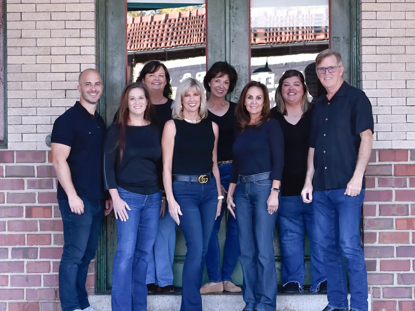 Group of eight adults standing together smiling in front of a green door with brick walls.