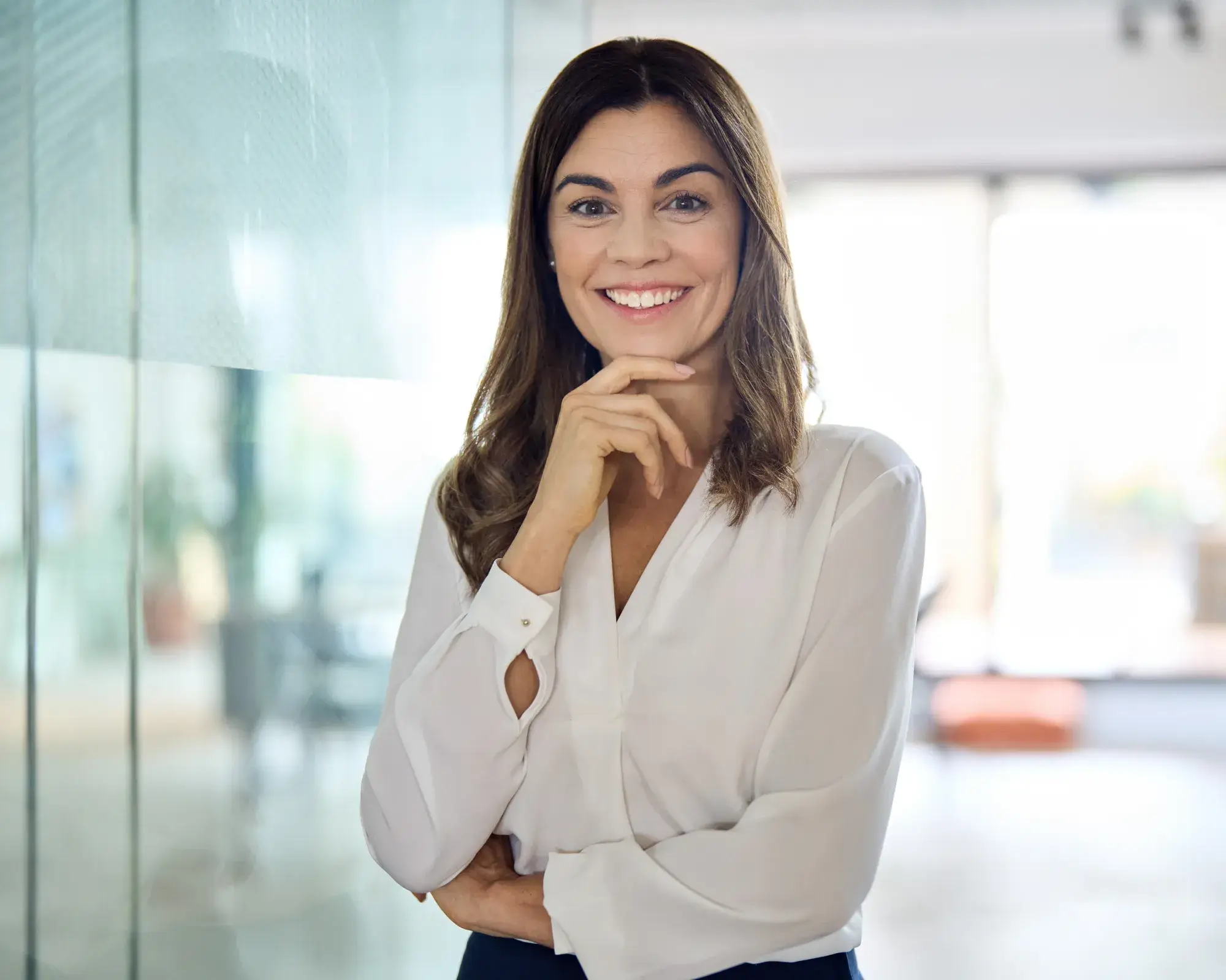 Smiling woman with long brown hair wearing a white blouse, standing confidently with hand on chin in a bright office.