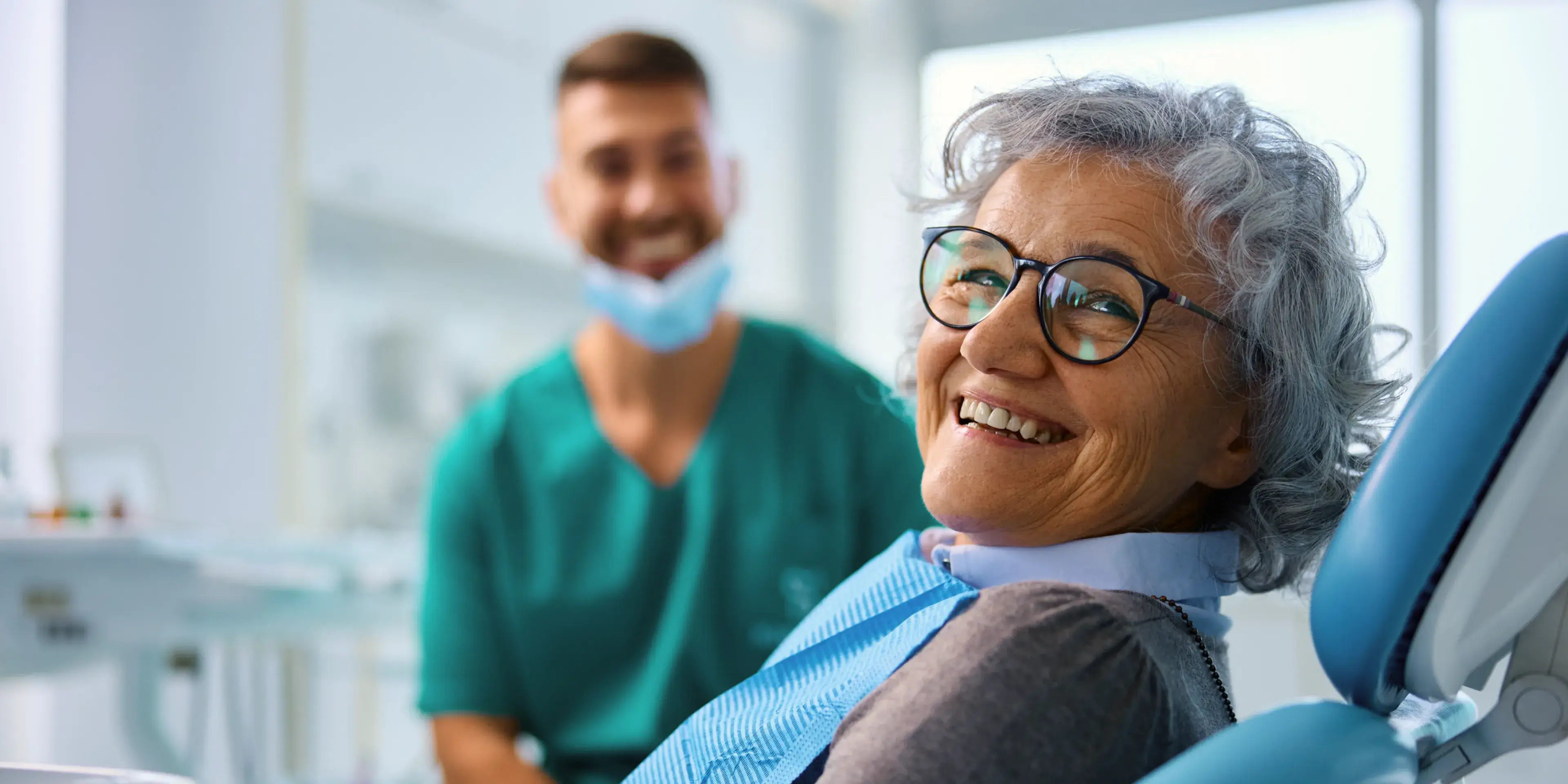 Smiling elderly woman wearing glasses sitting in a dental chair with a male dentist in a green uniform and mask in the background.