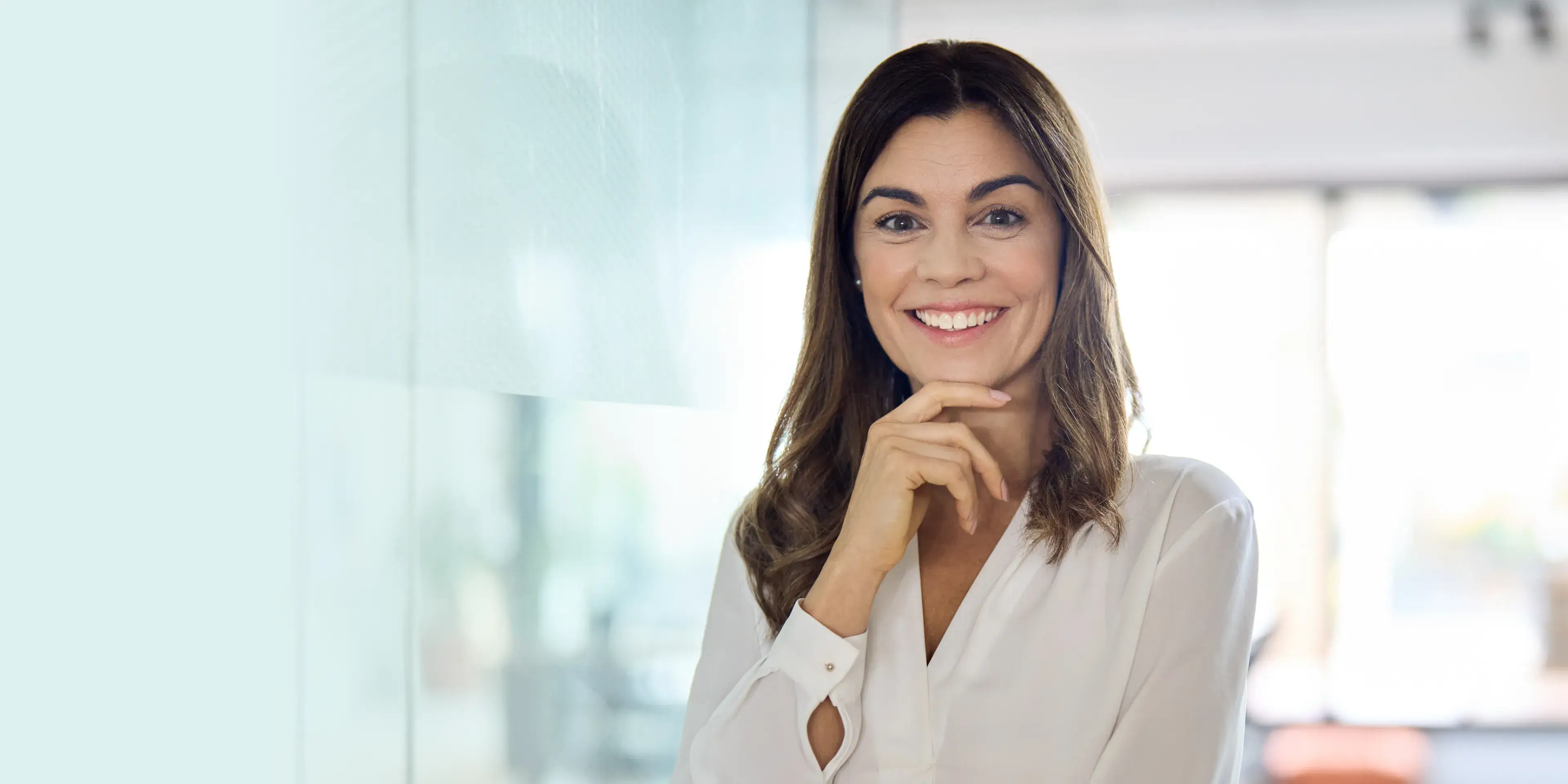 Smiling woman with long brown hair wearing a white blouse, standing indoors with one hand near her chin.