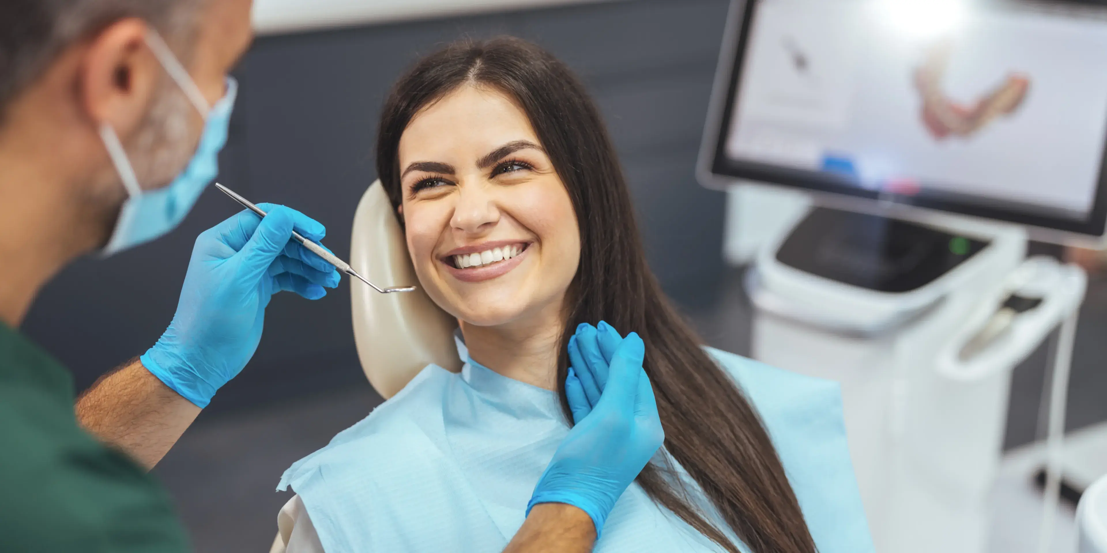Dentist wearing blue gloves and mask examining smiling woman's teeth in dental clinic.
