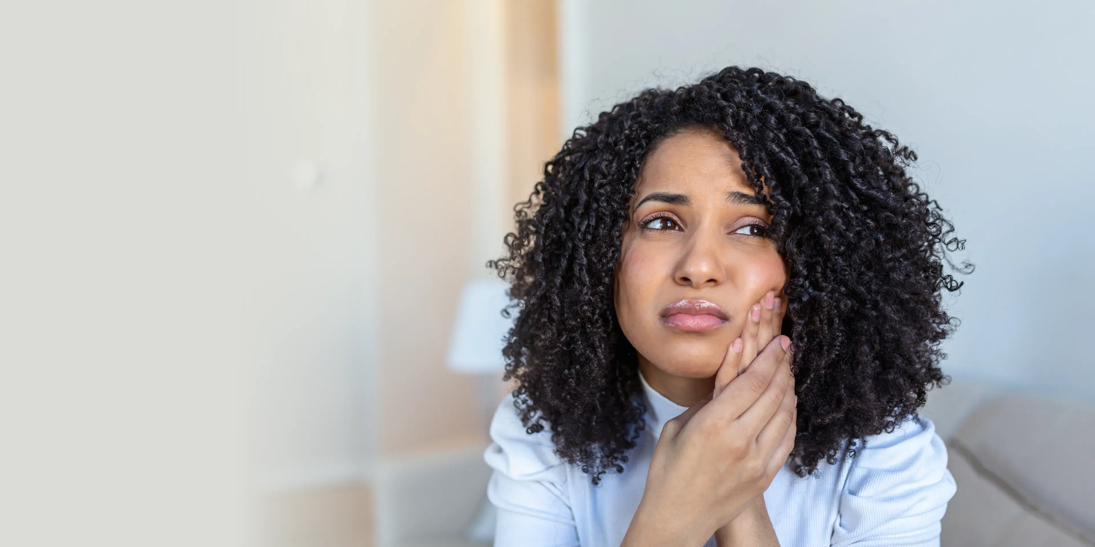 Young woman with curly hair holding her cheek and looking worried, indicating tooth pain.
